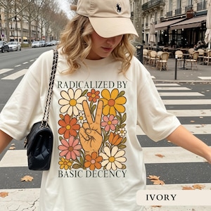 Peut inclure: T-shirt ivoire avec un motif rétro. Il présente un geste de la main en signe de paix entouré de fleurs orange, jaunes et roses. Le texte indique "RADICALIZED BY BASIC DECENCY". Le t-shirt est ample.