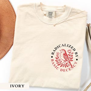 May include: An ivory short-sleeved t-shirt with a crew neckline. The chest features a circular graphic in red and black ink depicting a bird perched on a branch surrounded by flowers, with the text "RADICALIZED BY BASIC DECENCY" around it.