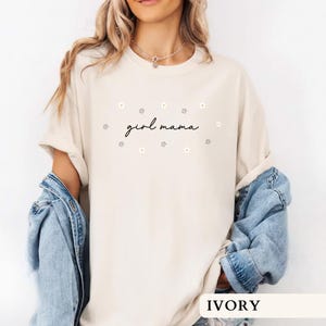 Op de afbeelding: Een ivoorkleurig T-shirt met korte mouwen en de tekst girl mama in een zwart cursief lettertype, omringd door kleine witte en gele madeliefjes. Het shirt heeft een ronde hals en een losse pasvorm, gecombineerd met een blauw spijkerjack.