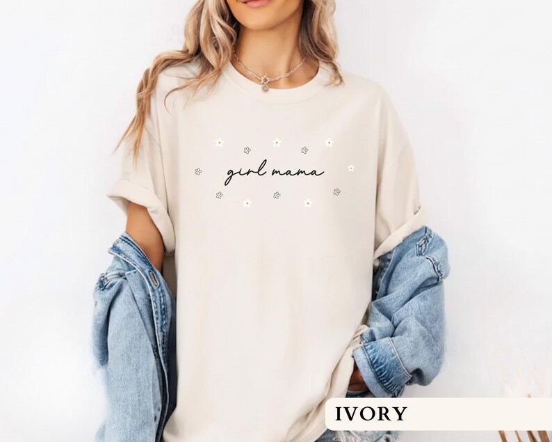 Op de afbeelding: Een ivoorkleurig T-shirt met korte mouwen en de tekst girl mama in een zwart cursief lettertype, omringd door kleine witte en gele madeliefjes. Het shirt heeft een ronde hals en een losse pasvorm, gecombineerd met een blauw spijkerjack.