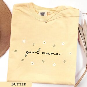 Op de afbeelding: Een botergeel T-shirt met korte mouwen en de zwarte cursieve tekst "girl mama" in het midden op de borst, omringd door kleine witte en zwart omlijnde bloemillustraties. Het shirt heeft een ronde hals en een Comfort Colors label.
