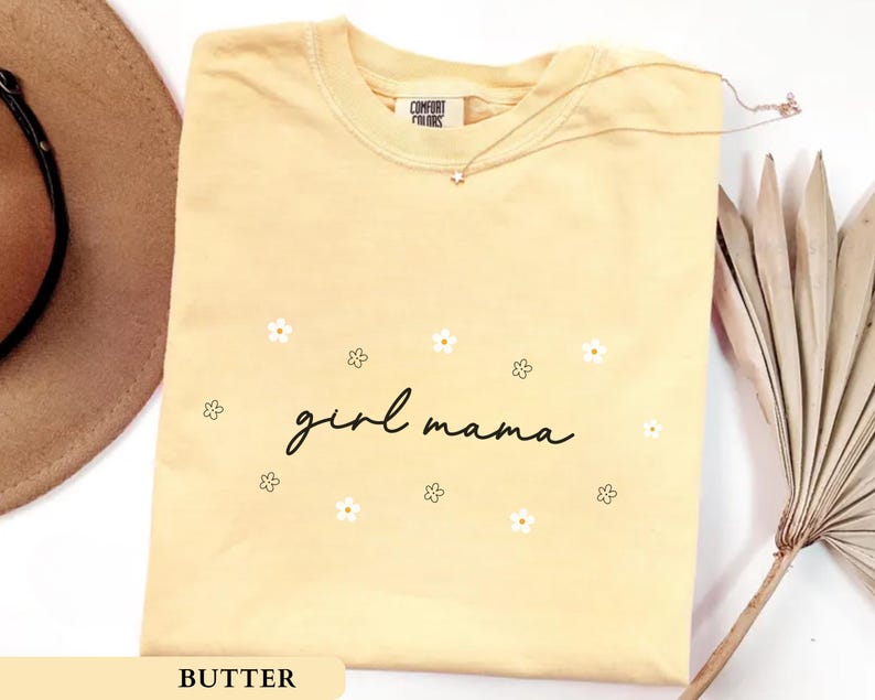Op de afbeelding: Een botergeel T-shirt met korte mouwen en de zwarte cursieve tekst "girl mama" in het midden op de borst, omringd door kleine witte en zwart omlijnde bloemillustraties. Het shirt heeft een ronde hals en een Comfort Colors label.