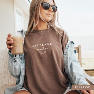 May include: A woman wearing a brown t-shirt with the text "AIRPLANE MODE" and a small airplane graphic. She is holding a glass of iced coffee and wearing a denim jacket.