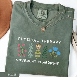 Op de afbeelding: Een mosgroen t-shirt met de woorden "PHYSICAL THERAPY" en "MOVEMENT IS MEDICINE" in het wit gedrukt. Het shirt heeft een bloemenontwerp in roze, geel en blauw. Een gouden ketting met een hanger ligt op het shirt.
