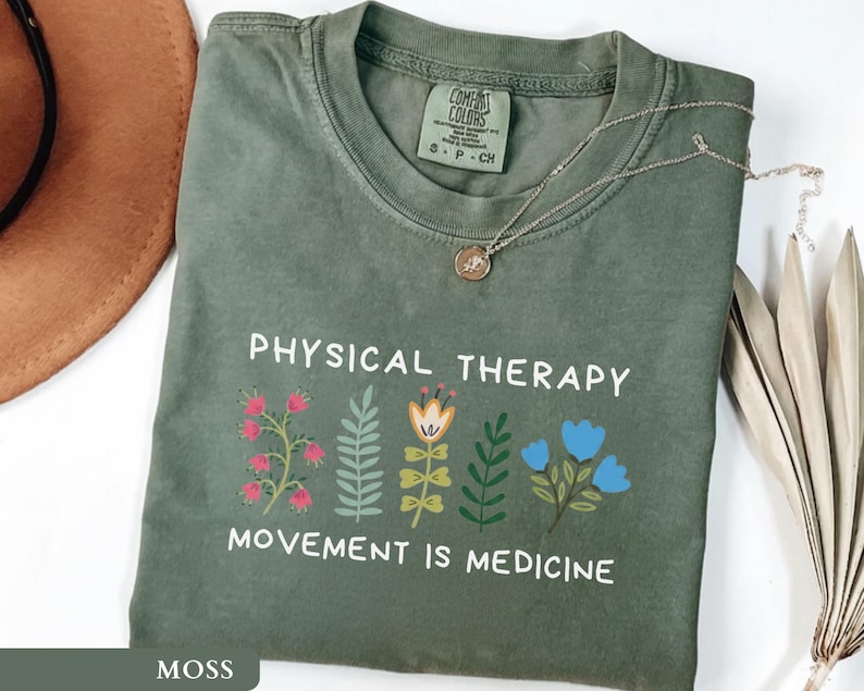 Op de afbeelding: Een mosgroen t-shirt met de woorden "PHYSICAL THERAPY" en "MOVEMENT IS MEDICINE" in het wit gedrukt. Het shirt heeft een bloemenontwerp in roze, geel en blauw. Een gouden ketting met een hanger ligt op het shirt.