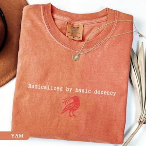 May include: A coral-colored t-shirt with the text "Radicalized by basic decency" and a red bird graphic. The shirt has a crew neck and a small tag. A gold necklace and a brown hat are also visible.