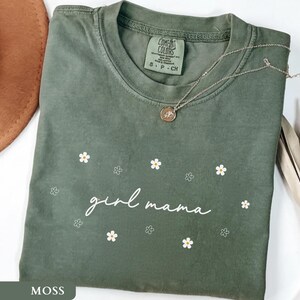 Op de afbeelding: Een mosgroen t-shirt met korte mouwen en de witte cursieve tekst girl mama in het midden op de borst, omringd door kleine witte en gele madeliefjesafbeeldingen. Het shirt heeft een ronde hals en een label van het merk Comfort Colors.