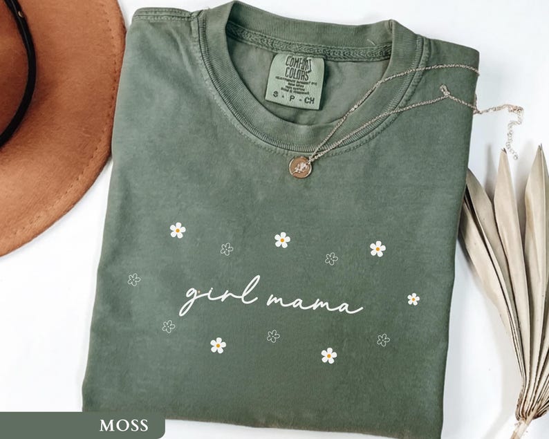 Op de afbeelding: Een mosgroen t-shirt met korte mouwen en de witte cursieve tekst girl mama in het midden op de borst, omringd door kleine witte en gele madeliefjesafbeeldingen. Het shirt heeft een ronde hals en een label van het merk Comfort Colors.