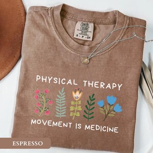 Op de afbeelding: Espresso-kleurig T-shirt met de witte tekst "PHYSICAL THERAPY MOVEMENT IS MEDICINE". Het shirt heeft kleurrijke bloemenillustraties. Een zilveren ketting met een hanger ligt op het shirt.