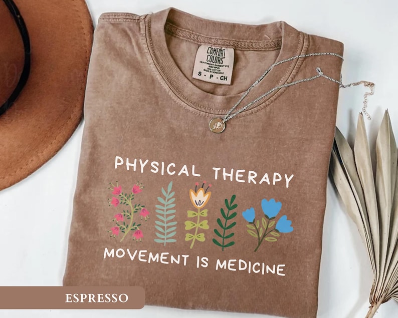 Op de afbeelding: Espresso-kleurig T-shirt met de witte tekst "PHYSICAL THERAPY MOVEMENT IS MEDICINE". Het shirt heeft kleurrijke bloemenillustraties. Een zilveren ketting met een hanger ligt op het shirt.