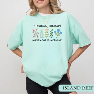 Op de afbeelding: Licht turquoise t-shirt met de zwarte tekst "PHYSICAL THERAPY" en "MOVEMENT IS MEDICINE". Het shirt heeft bloemenillustraties in roze, groen en blauw. De rechter benedenhoek van de afbeelding heeft de tekst "ISLAND REEF".