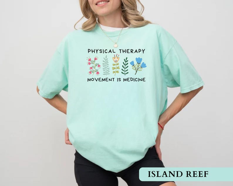 Op de afbeelding: Licht turquoise t-shirt met de zwarte tekst "PHYSICAL THERAPY" en "MOVEMENT IS MEDICINE". Het shirt heeft bloemenillustraties in roze, groen en blauw. De rechter benedenhoek van de afbeelding heeft de tekst "ISLAND REEF".