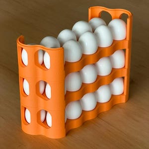 Pode incluir: Um suporte de plástico laranja para ovos cheio de ovos brancos. O suporte tem um design em camadas com bordas curvas e detalhes recortados nas laterais. Os ovos estão cuidadosamente dispostos em fileiras.