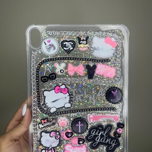 May include: A clear phone case with a silver glitter background and various black and pink Hello Kitty themed embellishments. The case has a black chain border and includes the text "girl gang" and "sweet".