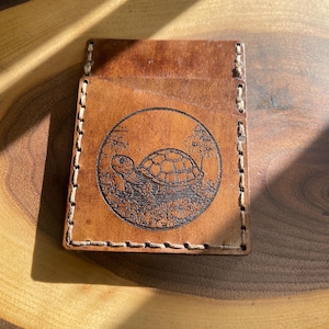 Könnte beinhalten: Eine braune Lederbörse mit einem genähten Rand und einem runden Design einer Schildkröte in einem Teich. Die Schildkröte ist schwarz umrandet und der Teich ist mit Blumen gefüllt.