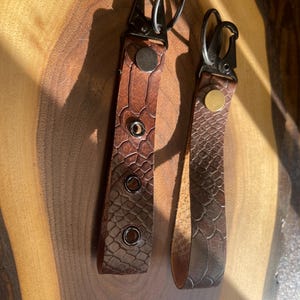 May include: Two brown leather keychains with metal rings and clasps. One keychain has four metal eyelets.