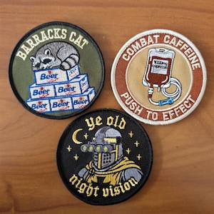 May include: Three circular embroidered patches. The top left is olive green with a raccoon on beer boxes and text BARRACKS CAT. The top right is tan with an IV bag and text COMBAT CAFFEINE PUSH TO EFFECT. The bottom is black with a knight wearing night vision goggles and text YE OLD NIGHT VISION.