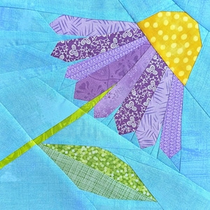 May include: A fabric flower quilt block with a yellow polka-dotted center, purple petals, a green stem, and a green and white leaf. The background is a light blue fabric.