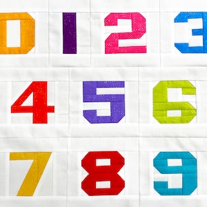 Puede incluir: Un fondo de tela blanco con nueve cuadrados de fieltro de colores, cada uno con un número del 0 al 9. Los números son de diferentes colores: amarillo, morado, rosa, azul, rojo, verde, amarillo, rojo y azul.