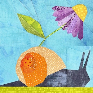 Op de afbeelding: Een kleurrijk patchwork-ontwerp met een slak en een bloem. De slak heeft een oranje huisje met witte stippen. De bloem heeft paarse bloemblaadjes en een gele kern. De achtergrond is lichtblauw met groene accenten.