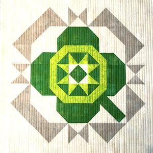 May include: A geometric quilt featuring a central green and yellow design resembling a four-leaf clover. The clover is surrounded by a gray and white geometric pattern. The quilt is made of fabric with a quilted texture.