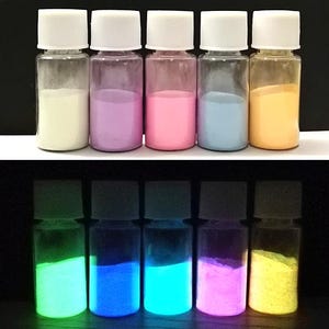 May include: Six small glass bottles with white lids containing different colored glow-in-the-dark powders. The powders are white, purple, pink, blue, and orange in the top row and green, blue, green, pink, and yellow in the bottom row.