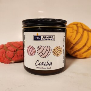 May include: A soy wax candle in a dark jar with a black lid. The label features the text "Concha (Mexican Sweet Bread)" and three heart-shaped designs. The candle is 13 oz (370 g). Two concha pastries are in the background.