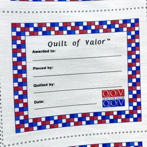 May include: A white fabric with a printed design featuring a central rectangular area with the text "Quilt of Valor". The border is a pattern of red, white, and blue squares. The text includes lines for filling in details.