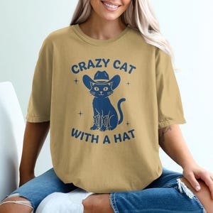 Puede incluir: Camiseta color mostaza con un gráfico azul de un gato con sombrero y botas de vaquero. El texto "CRAZY CAT WITH A HAT" está encima y debajo de la ilustración del gato. La camiseta tiene un corte holgado.