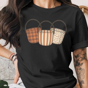 May include: Black t-shirt featuring a graphic of three decorative baskets with brown handles. The baskets have different patterns: orange plaid, vertical stripes, and a dotted design. The overall theme is autumn or harvest.
