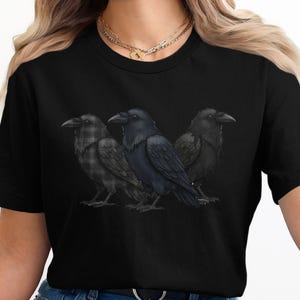May include: Black t-shirt with a graphic of three ravens. The central raven is solid black, with ravens in plaid and textured patterns on either side. The ravens are realistically illustrated.