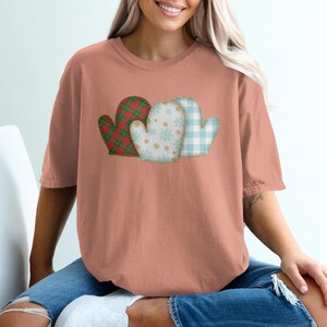 Puede incluir: Camiseta de color coral con tres diseños de mitones. Los mitones tienen un estilo patchwork con cuadros rojos y verdes, blancos con copos de nieve y estampados de cuadros vichy azul claro. La camiseta es de estilo casual y cómodo.