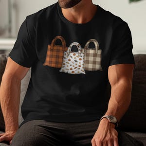 Può includere: T-shirt nero con tre borse. Le borse presentano diversi motivi: quadri arancioni e marroni, bianco con zucche e caramelle, e quadri marroni e beige. Le borse hanno manici marroni.