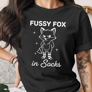 May include: Black t-shirt with the text "FUSSY FOX in Socks" in white. The graphic is a cartoon fox with an angry expression, wearing striped socks. The fox is outlined in white.