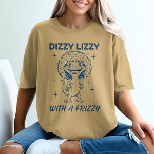 May include: Mustard-coloured t-shirt with a cartoon lizard sporting a curly afro. The text "DIZZY LIZZY" is above the lizard, and "WITH A FRIZZY" is below. The design is in navy blue.