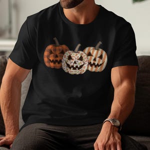 May include: Black t-shirt with a design of three Halloween pumpkins. The pumpkins have carved faces and different patterns: orange plaid, floral, and orange and white stripes. A casual, festive garment for autumn.