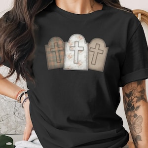 May include: Black t-shirt featuring three tombstone designs with crosses. The tombstones have different patterns: plaid, floral, and solid. The crosses are outlined in a contrasting color, creating a layered effect. The shirt is a casual wear item.