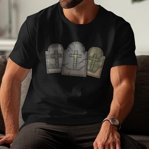 May include: Black t-shirt featuring a graphic of three tombstones with crosses. The tombstones are in shades of gray and green, with a cracked texture. The design is centered on the shirt.