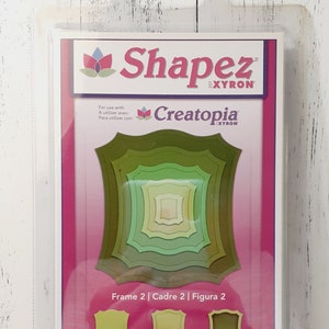 May include: A package containing a set of five green nested frame dies. The packaging features the "Shapez" and "Creatopia" logos, along with instructions in English, French, and Spanish. The package also includes the words "Cut!", "Emboss!", and "Stencil!"