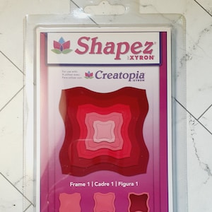 May include: A package of five Spellbinders Shapez Creatopia dies for use with die-cutting machines. The dies are red, pink, and white and feature a square frame with rounded corners. The package includes the text "Frame 1 | Cadre 1 | Figura 1", "Cut! Couper! ¡Cortar!", "Emboss! Embosser! ¡Estampar!", "Stencil! Pochoir! ¡Estarcir!" and "Spellbinders 5 Dies Included Formes comprises Troqueles incluido".