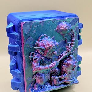 May include: A rectangular blue storage case featuring a 3D-printed relief on the front panel. The relief depicts a goblin-like figure with pointed ears and goggles, interacting with a mechanical device, rendered in a shifting iridescent purple, green, and blue finish.