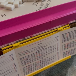 May include: A Mah Jongg game set featuring a white board with a pink plastic frame, a pile of white rectangular tiles, and a yellow plastic tile rack. A printed score card with black text and columns of values is held by the yellow rack.