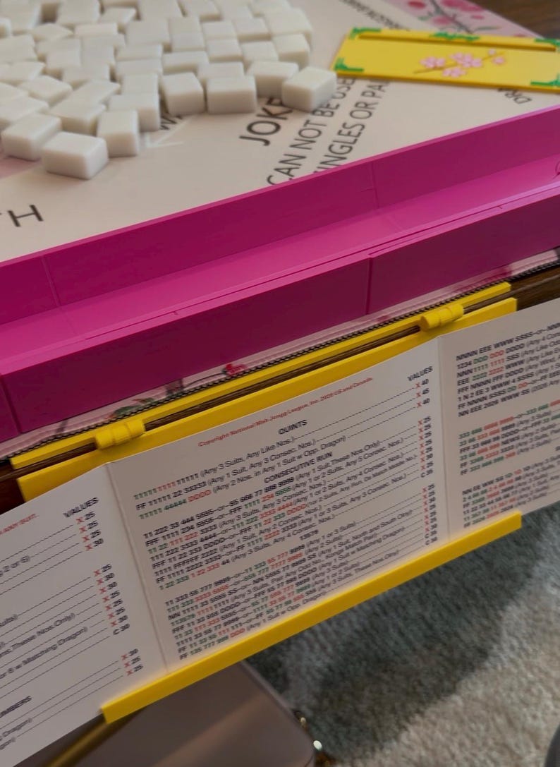 May include: A Mah Jongg game set featuring a white board with a pink plastic frame, a pile of white rectangular tiles, and a yellow plastic tile rack. A printed score card with black text and columns of values is held by the yellow rack.
