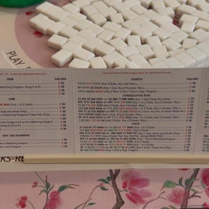 May include: A Mah Jongg score card featuring columns of game rules, point values, and hand combinations printed in black and red text on white paper, held upright by a thin beige plastic stand, with white game tiles visible in the background.