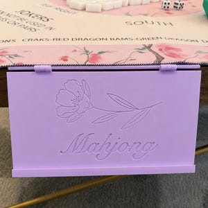Könnte beinhalten: Ein hellvioletter, rechteckiger Mahjong-Fliesenhalter mit einer klappbaren Oberkante und einem eingeprägten Blumenmuster über dem Wort Mahjong in Schreibschrift. Der Halter ist vertikal an der Kante eines Tisches mit einer Spielmatte angebracht.