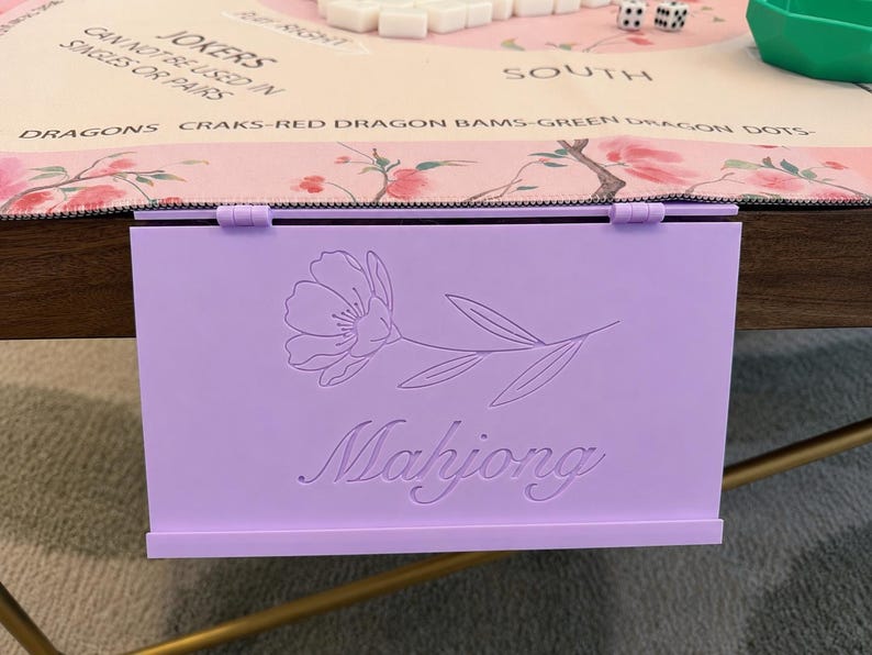 May include: A light purple rectangular Mahjong tile rack featuring a hinged top edge and a debossed floral design above the word Mahjong in a cursive script. The rack is positioned vertically against the edge of a table with a game mat.