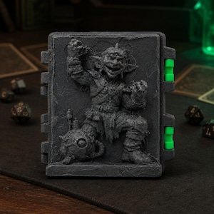 May include: A rectangular dark gray storage box featuring a relief sculpture of a goblin on the front cover. The box has two bright green hinges on the right side and a textured, stone-like finish across the entire surface of the container.