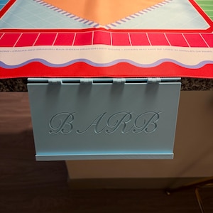 May include: A light blue plastic card holder with the name BARB engraved in a cursive font on the front, attached to a colorful Mah Jongg mat featuring a central peach diamond, blue grid patterns, and red borders with printed game rules.