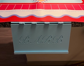 Puede incluir: Un tarjetero de plástico azul claro con el nombre BARB grabado en letra cursiva en el frente, sujeto a un tapete de Mah Jongg colorido con un diamante central color melocotón, patrones de cuadrícula azules y bordes rojos con reglas impresas.