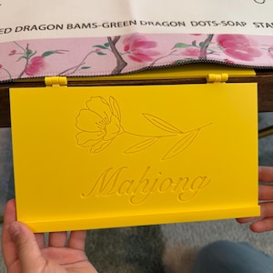 May include: A bright yellow rectangular Mahjong tile rack featuring a hinged design. The front surface is engraved with a delicate line-art illustration of a single flower with leaves and the word Mahjong written in a cursive script font.